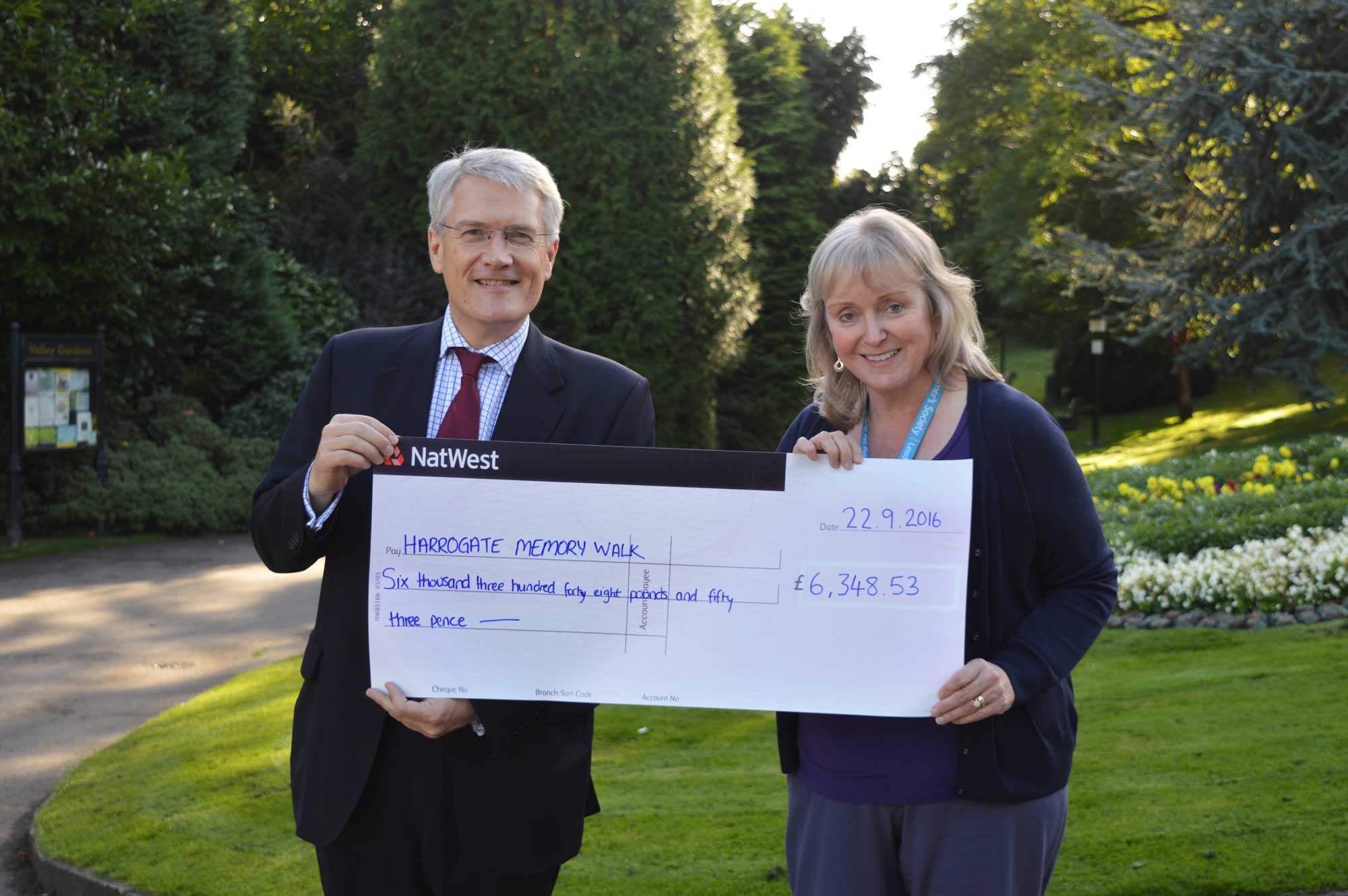 Andrew Jones MP hands a cheque for £6,348.53 to Alzheimer’s Society Services Manager, Alison Wrigglesworth at the entrance to the Valley Gardens where the walk took place