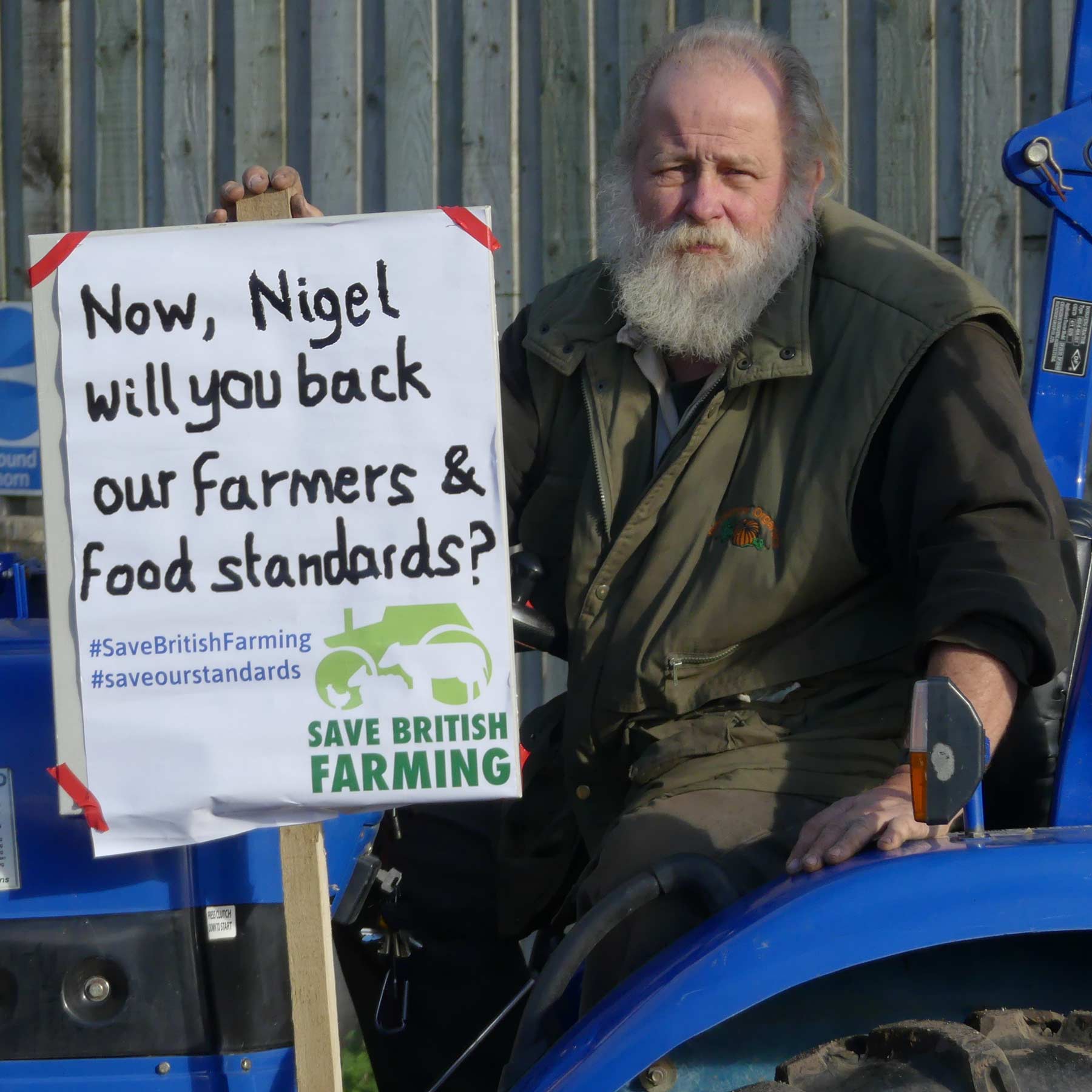 Arnold Warneken sitting on his blue tractor, holding a poster on his right saying ‘Now Nigel will you back our farmers and food standards?’