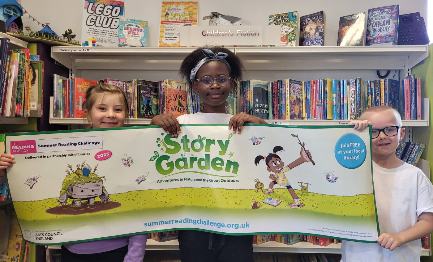 From left, Rita, Sharon and Lotan from Bentham Community Primary School, showing off the Story Garden Summer Reading Challenge banner